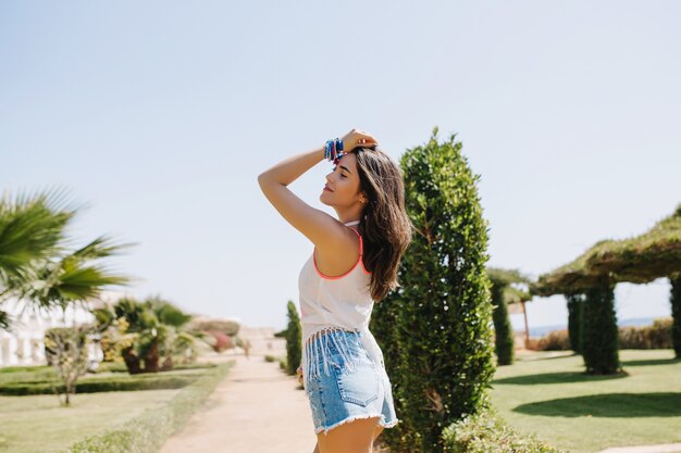 Knap vrolijk meisje in vintage tank-top poseren buiten in zonnige Zomerochtend, genietend van weekend. Schattige vrolijke vrouw in stijlvolle accessoires wandelen door het steegje in het exotische park