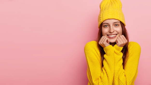 Knap vrouwelijk model houdt kin met beide handen vast, glimlacht zachtjes naar de camera, gekleed in een stijlvolle gele hoed en trui, modellen over roze muur.