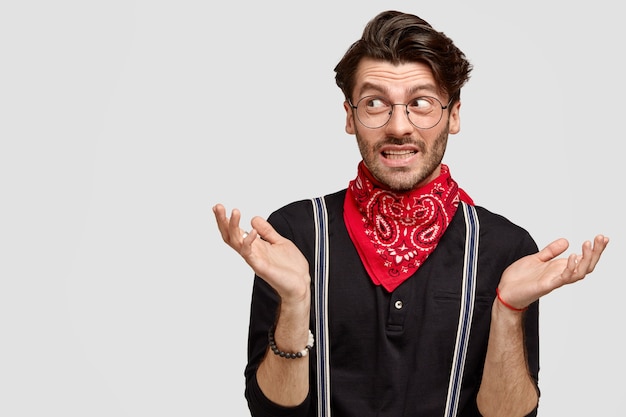 Knappe aarzelende man haalt zijn schouders op, kijkt twijfelend opzij, weet niet wat hij moet zeggen, draagt een stijlvol shirt en een rode bandana om de nek