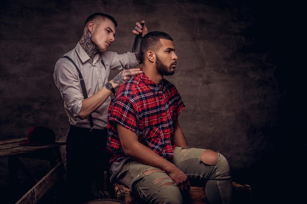 Knappe Afro-Amerikaanse man die een kapsel krijgt terwijl hij op houten kisten in een studio zit. Ouderwetse professionele getatoeëerde kapper doet een kapsel.