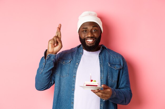 Knappe Afro-Amerikaanse man die verjaardag viert, wens doet met gekruiste vingers, verjaardagstaart met kaars vasthoudt, staande tegen roze achtergrond