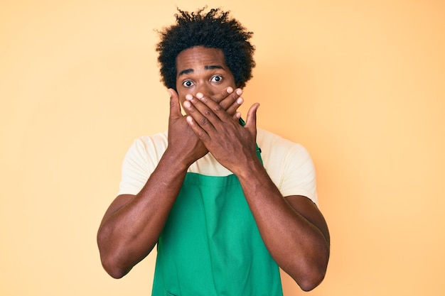 Knappe afro-amerikaanse man met afrohaar die oberschort draagt geschokt mond bedekkend met handen voor fout. geheim concept.