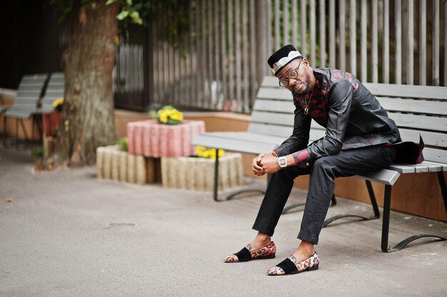 Knappe afro-amerikaanse man met traditionele kledingpet en bril in moderne stad zittend op de bank