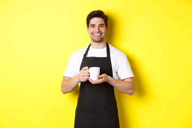 Knappe barista die koffie serveert, beker meeneemt, in zwart schort staat met een vriendelijke glimlach.