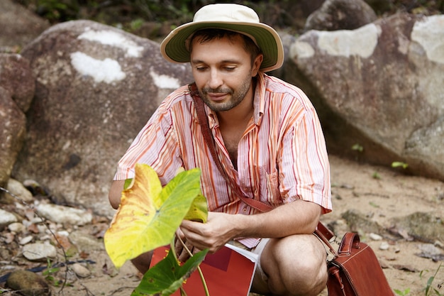 Knappe bebaarde bioloog met hoed die blad van groene plant houdt, kijkt met vriendelijke en zorgzame uitdrukking tijdens zijn milieustudies op het werkveld.