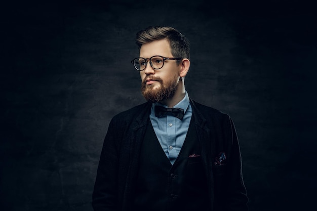 Knappe bebaarde elegante man in bril gekleed in een donkerblauw pak met vlinderdas over grijze vignet achtergrond.