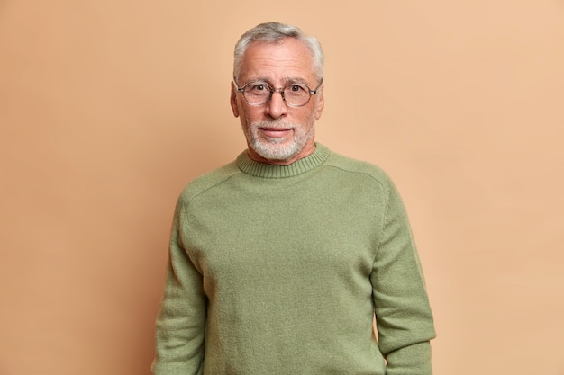 Knappe bebaarde Europese man met nieuwsgierige blik draagt een bril en basic jumper kijkt direct front poses tegen beige muur