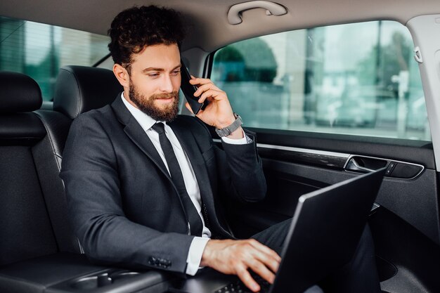 Knappe, bebaarde, glimlachende zakenman die op zijn laptop werkt en een mobiele telefoon spreekt op de achterbank van de auto