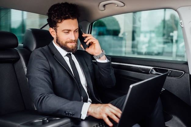 Knappe, bebaarde, glimlachende zakenman die op zijn laptop werkt en een mobiele telefoon spreekt op de achterbank van de auto