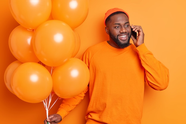 Knappe bebaarde man heeft vrolijk gesprek via smartphone viert positieve gebeurtenis nodigt vrienden op feestje versiert kamer met ballonnen geïsoleerd over oranje muur