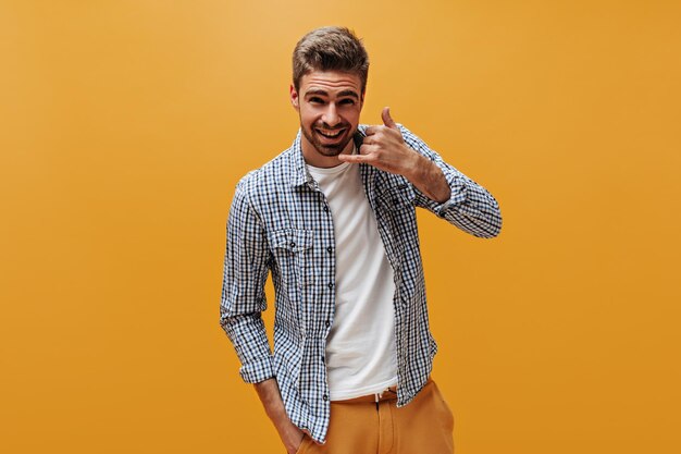 Knappe bebaarde man in lichte broek, wit t-shirt en geruit hemd glimlacht, kijkt in de camera en toont roepnaam op oranje achtergrond