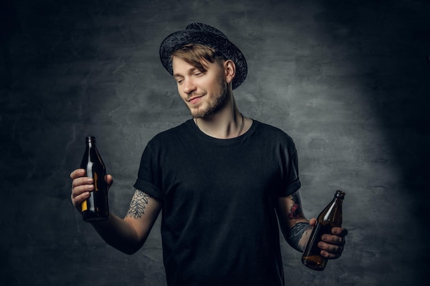 Gratis foto knappe bebaarde man met getatoeëerde armen, gekleed in een zwart t-shirt en hoge hoed houdt ambachtelijk gebotteld bier vast.