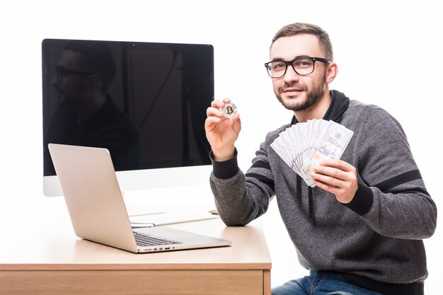 Knappe bebaarde man op zijn werkplek met laptop en pc-beeldscherm achterop met bitcoin en dollars contant in handen