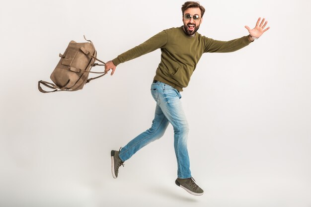Knappe bebaarde stijlvolle man springen uitgevoerd geïsoleerd gekleed in sweatshirt met reistas, spijkerbroek en zonnebril, gekke reiziger op haast