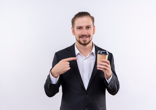 Knappe bedrijfsmens die de koffiekop van de kostuumholding houdt die met vinger aan het glimlachen richtend zich over witte achtergrond bevindt