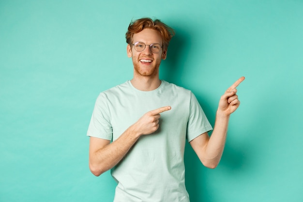 Knappe blanke man met gemberhaar, bril en t-shirt, wijzende vingers naar rechts en vrolijk lachend, reclame tonend, staande over turkooizen achtergrond.