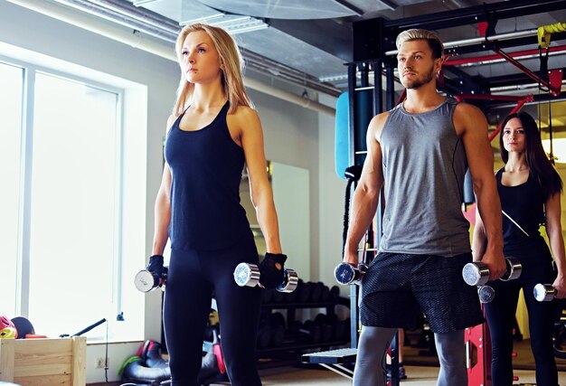 Knappe blonde, atletische man en twee slanke vrouwelijke fitnessmodellen die schouderoefeningen doen met halters in een sportschoolclub.