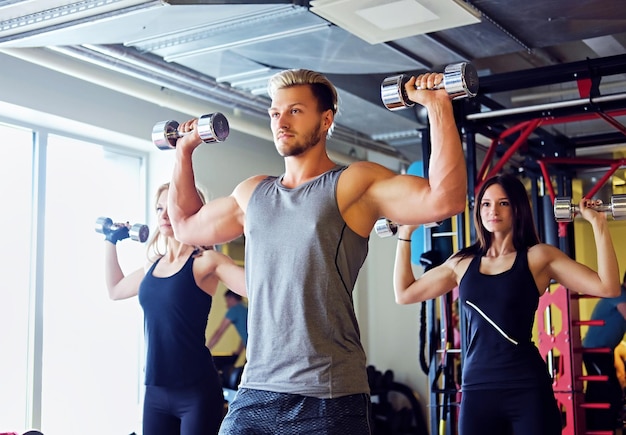 Knappe blonde, atletische man en twee slanke vrouwelijke fitnessmodellen die schouderoefeningen doen met halters in een sportschoolclub.