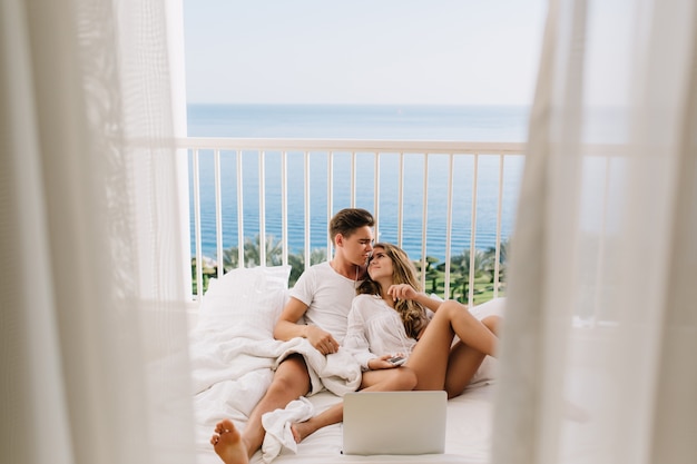 Knappe brunette man omhelst zijn mooie vrouw, liggend op bed met witte laptop. Mooie jonge paar tijd samen doorbrengen in luie ochtend, kussen op balkon met uitzicht op zee