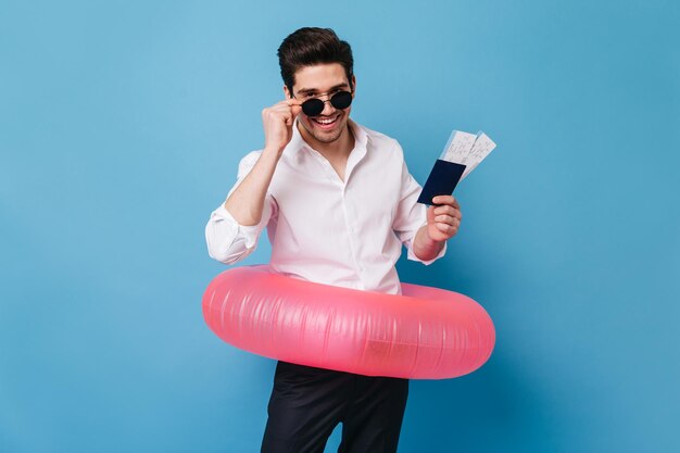 Knappe brunette man zet zijn bril af en kijkt naar de camera met een glimlach op blauwe achtergrond Guy in wit overhemd wacht op vakantie en poseren met kaartjes en opblaasbare cirkel