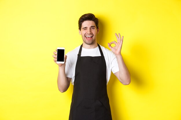 Knappe coffeeshopmedewerker die ok teken en smartphonescherm toont, toepassing aanbeveelt, die zich over gele achtergrond bevindt.