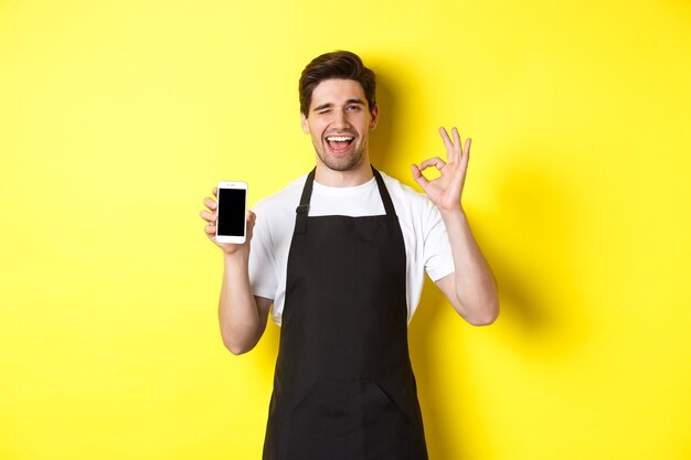 Knappe coffeeshopmedewerker die ok teken en smartphonescherm toont, toepassing aanbeveelt, staande over gele achtergrond