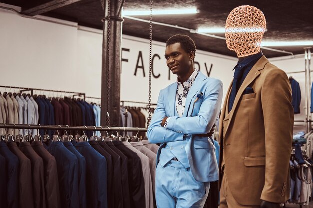 Knappe elegant geklede Afro-Amerikaanse man poseren met gekruiste armen in de buurt van mannequin in een klassieke herenkledingwinkel.