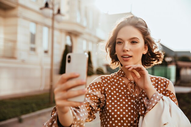 Knappe elegante vrouw met smartphone en selfie maken. Geweldige Europese dame in bruine jurk die zich voordeed op straat.