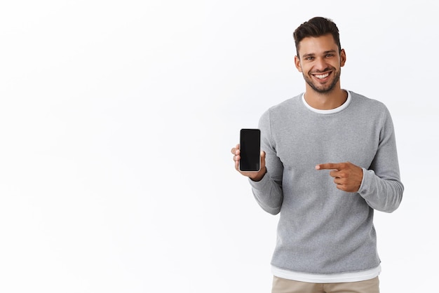 Knappe enthousiaste Glimlachende man met haren in grijze trui met smartphone scherm naar camera gericht mobiel display en grijnzend aanbevelen applicatie autodelen of winkelen site