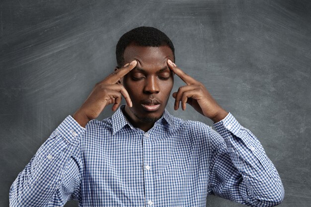 Knappe ernstige verbaasde Afrikaanse student gekleed in geruit overhemd zijn voorhoofd beroven, zijn ogen sluiten, geconcentreerd en gefocust kijken, proberen het juiste antwoord te onthouden tijdens de test in de klas
