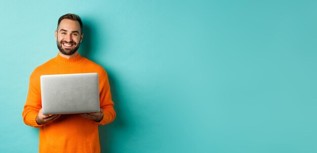Knappe freelancer die aan laptop werkt en glimlachend in oranje trui over lichtblauwe backgro staat