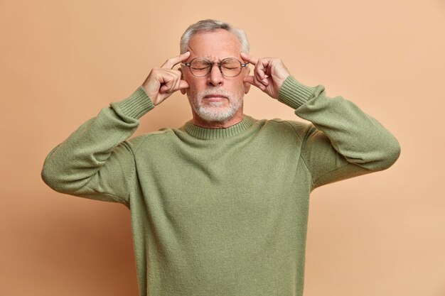 Knappe gefrustreerde man heeft vreselijke migraine houdt de handen op de slapen, sluit de ogen om pijn te onthullen staat moe draagt een bril en trui geïsoleerd over bruine muur