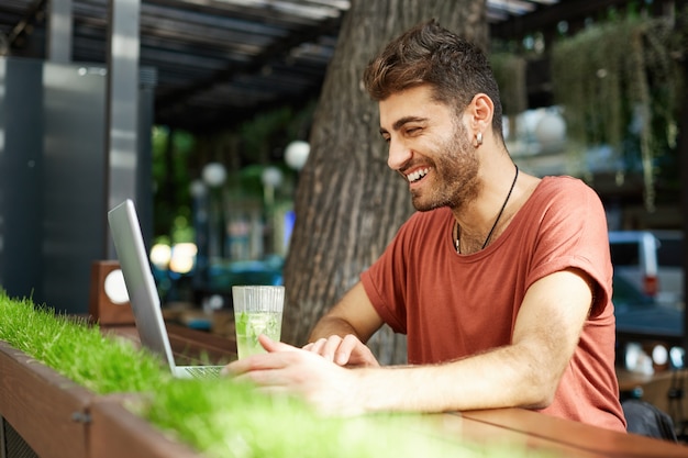 Knappe gelukkig bebaarde man in oortelefoons met laptop op terras, vrolijk glimlachend