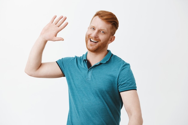 Knappe gelukkig roodharige man wacht op high five, glimlachend breed palm hoog opheffen om persoon op moderne manier te begroeten, close over grijze muur verwelkomen, trendy poloshirt dragen