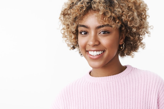 Knappe gelukkige Afro-Amerikaanse vrouwelijke student met afro blond kapsel in trui met vrolijke brede glimlach tevreden naar de camera staren met zorgeloze uitdrukking plezier, gelukkig lachen