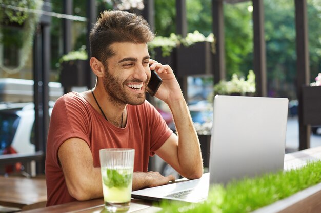 Knappe Glimlachende man praten op de mobiele telefoon, de verkoper bellen die hij online vond tijdens het gebruik van laptop, winkelen, appartement op internet zoeken