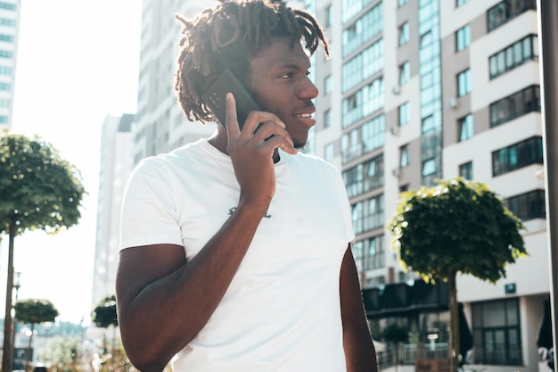 Knappe hipster modelOngeschoren Afrikaanse man gekleed in witte zomer tshirtMode man met dreadlocks kapsel Poseren in de straatGebruik heeft smartphone Praten met vrienden op mobiel