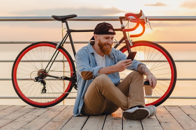 Knappe hipster stijl bebaarde man dragen denim shirt en pet smartphone met fiets houden in de ochtend zonsopgang door de zee koffie drinken, gezonde actieve levensstijl reiziger