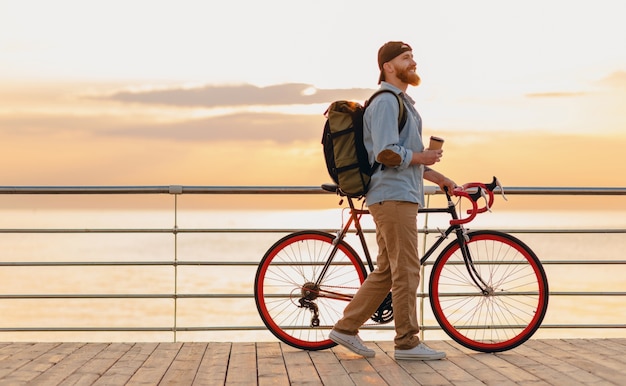 Knappe hipster stijl bebaarde man met rugzak denim shirt en pet met fiets dragen in de ochtend zonsopgang door de zee koffie drinken, gezonde actieve levensstijl reiziger backpacker