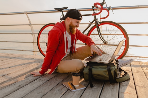 Gratis foto knappe hipster stijl bebaarde man online freelancer aan het werk op laptop met rugzak en fiets in de ochtend zonsopgang door de zee gezonde actieve levensstijl reiziger backpacker