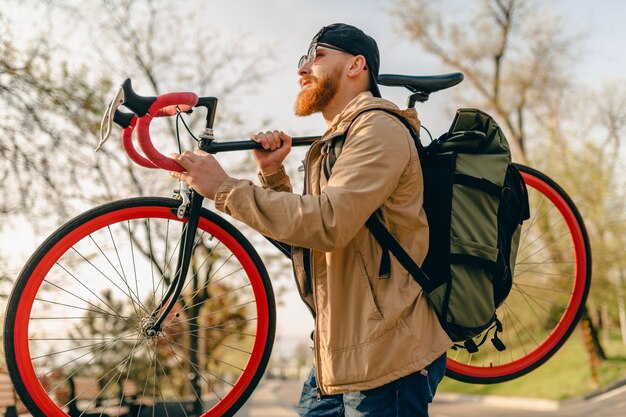 Knappe hipster stijlvolle bebaarde man in jas en zonnebril alleen lopen in straat met rugzak op fiets gezonde actieve levensstijl reiziger backpacker