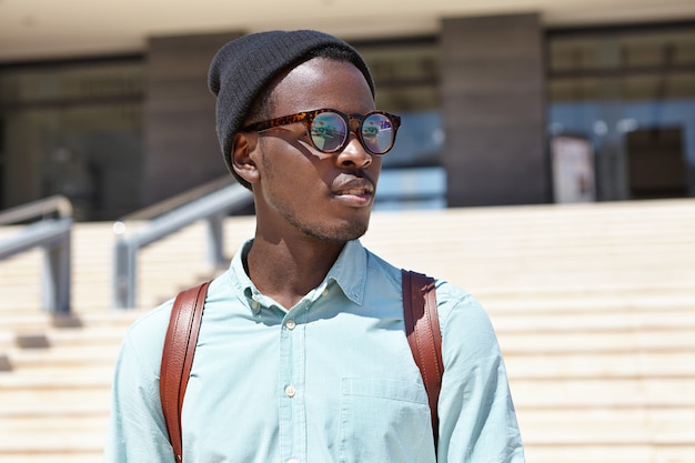 knappe jonge Afrikaanse mannelijke toerist die rugzak draagt die straten van onbekende buitenlandse stad verkent terwijl hij op vakantie in het buitenland, modern gebouw en betonnen trappen