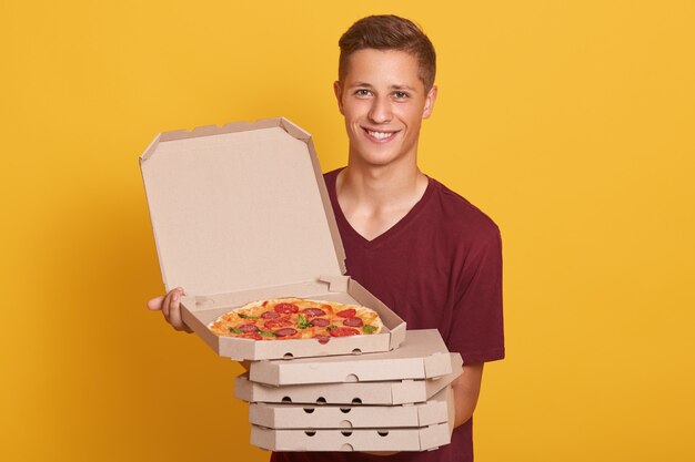 Knappe jonge bezorger met stapel pizzadozen, gekleed casual t-shirt, camera kijken en glimlachen, open doos met smakelijke pepperoni tonen, poseren geïsoleerd op gele studio