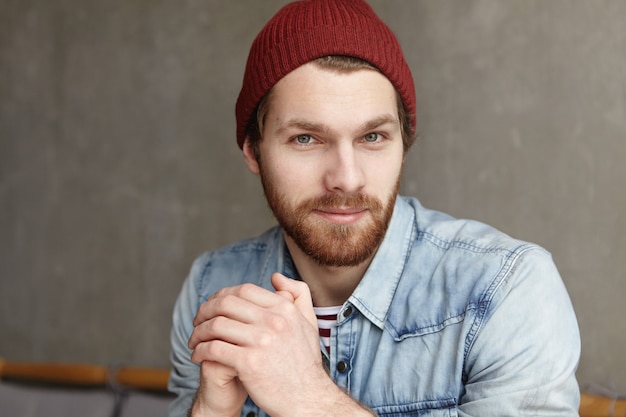 Knappe jonge blanke man met dikke baard op zoek met flirtende glimlach, handen gevouwen. aantrekkelijke man stijlvolle kleding dragen