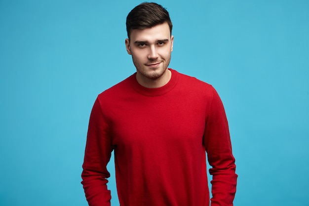 Knappe jonge donkerharige man stijlvolle rode trui dragen met lange mouwen glimlachend in de camera