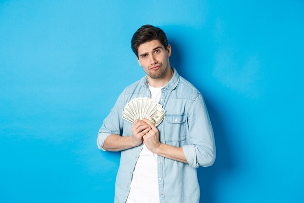 Knappe jonge man die geld voor zichzelf houdt, glimlachend en hebzuchtig kijkt, staande over een blauwe achtergrond.