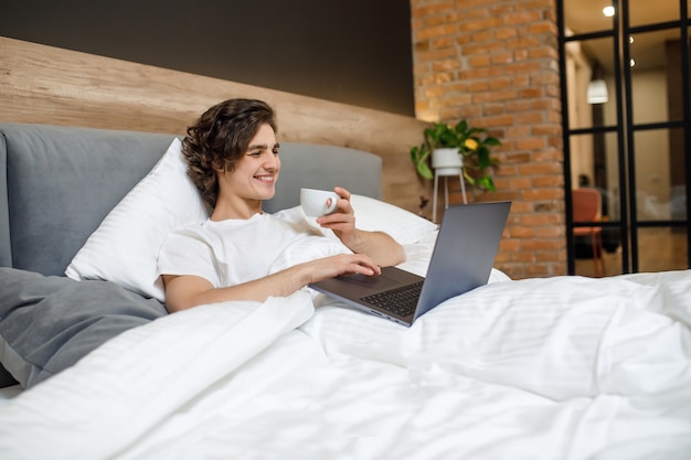 Knappe jonge man die 's ochtends op zijn bed ligt, een kopje koffie of thee vasthoudt en een laptop gebruikt