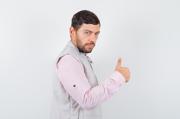 Knappe jonge man duim opdagen in shirt, vest en serieus kijken.