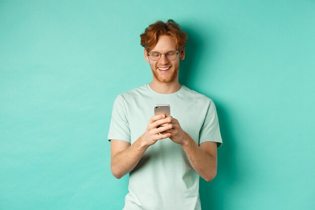 Knappe jonge man in glazen met rood slordig haar bericht lezen op mobiele telefoon, glimlachend en kijken naar scherm, staande op mint achtergrond.