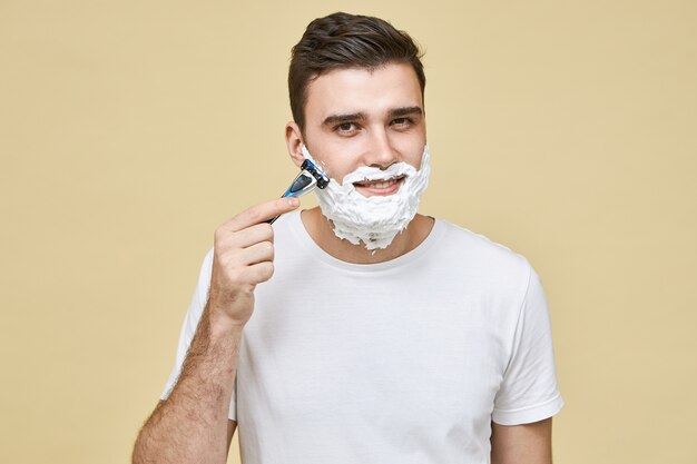 Knappe jonge man in wit t-shirt met scheermesje terwijl het scheren van baard tegen korrel om huidirritatie met glimlach te voorkomen, het verzorgen van zijn uiterlijk. Mannelijkheid, stijl en schoonheid